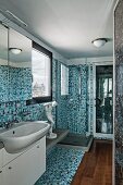 Schmales Bad gefliest mit Mosaikfliesen in verschiedenen Blautönen, weißer Waschtisch mit Spiegelschrank und Holzdielen vor Fliesenstreifen am Boden