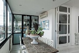 Bepflanzte Amphore auf rustikalem Holztisch und traditionelle weiß lackierte Stühle in Loggia ähnlichem Raum