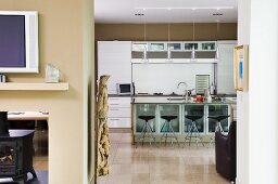 View into open-plan kitchen with sand-coloured walls, bar stools with black shell seats and counter with glass doors on base unit