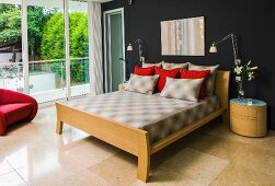 Scatter cushions on double bed with wooden frame and headboard, designer table lamp on oval bedside cabinet against black-painted wall, sand-coloured limestone floor, glass wall to one side