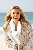 A young blonde woman by the sea wearing a beige knitted cardigan and a scarf