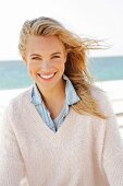 A young blonde woman by the sea wearing a denim shirt and a white woollen jumper