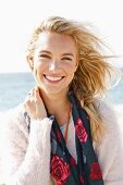 A young blonde woman by the sea wearing a light jumper and a scarf