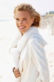 A young blonde woman on a beach wearing a white coat with a jumper over her shoulders