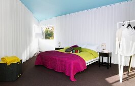 Triangular sleeping area screened by white curtains; double bed with bright bedspreads, dark carpet, standard lamp between windows and white clothes on clothes rack in foreground