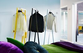 Shirts and jumpers on designer clothes racks behind partially visible bed with scatter cushions of various colours and green bedspread