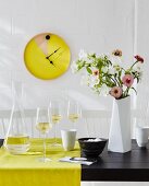 A home-made wall clock – an old tray painted and fitted with a clock mechanism on a whitewashed brick wall with a glass carafe and wine glasses on a yellow table runner next to a bunch of flowers in a white vase on a black table
