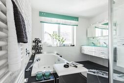 A modern, black and white bathroom with a bubble-jet bathtub and a chrome-plated laundry box stool in front of it with a double washstand in the background