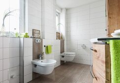 Raumhoch, weiss gefliestes Bad mit Bidet und Toilette an Fensterwand, auf Boden hellbraune Fliesen, teilweise hinter den Sanitärobjekten an Wand hochgezogen