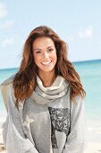 A young brunette woman by the sea wearing a grey sweatshirt and a scarf