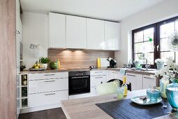 A white modern fitted kitchen with shiny white fronts and a wooden look dining table in the foreground