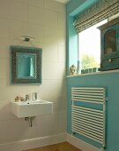 Spiegel mit marokkanischem Zierrahmen über modernem Waschbecken; türkisblaue Seitenwand mit Handtuch-Heizkörper und antikes Schränkchen auf Fensterbank