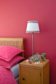 Bedside cabinet with lamp against raspberry wall next to Art Nouveau bed with cane headboard and pink bed linen