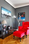 Bright red wing-back chair with footstool and reflections in glossy sideboard below surrealist painting on grey-painted wall