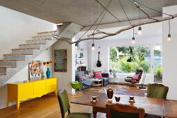 Lights hanging from large branch above oak table and upholstered chairs in open-plan interior; yellow sideboard below exposed concrete staircase