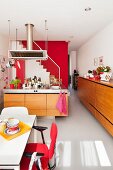 Dining area with red armchair, island kitchen counter, staircase against red wall and pale wood sideboard in open-plan interior