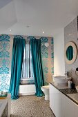 Fenster mit bodenlangem Vorhang in floral gemusterte Tapetenwand, seitlich ein Waschtisch mit Aufsatzbecken auf Unterschränken
