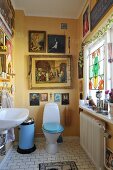Yellow-painted bathroom with toilet, paintings with religious motifs on wall and ornaments on windowsill to one side