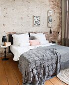 Graue, gehäkelte Tagesdecke auf Doppelbett vor teilweise verputzter Ziegelwand im Schlafzimmer mit Holzdielenboden