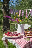 Torteletts und Waffeln mit frischen Kirschen und Wiesenblumen in weißem Krug auf Gartentisch mit karierter Tischdecke