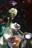 A breakfast tray in a garden with phlox flowers in a glass vase, croissants, a Thermos flask and a bowl of sugar