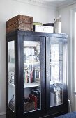 Books in glass-fronted cabinet with wooden crates and boxes on top on corner of room