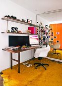 Weisser Schalenstuhl an schmalem Schreibtisch mit Wandborden und gelber Teppich vor verspiegelter Rückwand