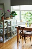 Delicate metal chair at rustic wooden table next to half-height, glass-fronted cabinet