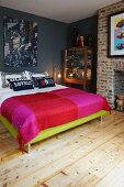 Double bed with blanket in different shades of red against grey-painted wall with fireplace in brick chimney breast to one side