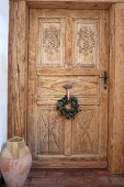 Wreath hung on carved interior door with rustic floor vase to one side