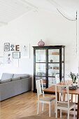 Dining area with white chairs, wooden table and glass-fronted cabinet next to grey sofa in open-plan interior