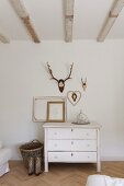 Antlers above white, vintage chest of drawers in bedroom with exposed ceiling beams