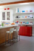 Island counter and white bar stools in open-plan kitchen with colourful crockery on white floating shelves above red chest of drawers