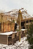 Stühle an langer Tafel unter Pergola aus Geflecht auf Baumstämmen in winterlichem Innenhof