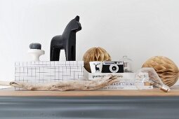 Still-life arrangement of black horse ornament on white box with pattern, vintage camera on stacked books and paper pompoms