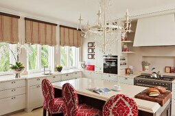 White country-house kitchen with free-standing counter and bar stools upholstered in red and white brocade below white chandelier