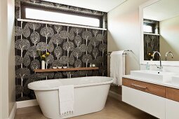 Free-standing bathtub against wall with transom window and black and white, tree-patterned wallpaper in designer bathroom
