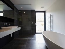 Schwarz-weisses Designerbad mit freistehender Badewanne auf dunklem Fliesenboden, gegenüber weisser Waschtisch und Dusche mit Glastrennwand