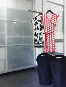 Moderner Waschraum mit schwarzen Wäschebehältern unter Wäscheständer, darauf Damenkleider gehängt, vor Schrank mit Glas Schiebetüren