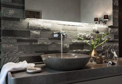 An illuminated mirror above polished limestone washstand with a console basin and grey limestone relief tiles