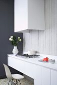 Minimalistische Küchenzeile mit Klassikerstuhl vor einem Gaskochfeld und verkleideter Dunstabzugshaube auf Sichtbetonwand