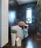 House-shaped lantern and floor vase in front of designer bathtub in dark-tiled bathroom; cat jumping down from edge of bath