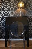 Ghost chair in front of black, antique bureau with table lamp against black wallpaper with ornamental silver pattern