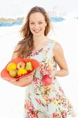 A young woman wearing a summer dress holding a hat filled with fruit and holding out an apple