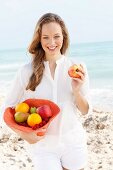 A young woman by the sea wearing a white blouse and shorts holding a hat filled with fruit