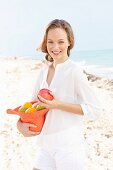 A young woman by the sea wearing a white blouse and shorts holding a hat filled with fruit