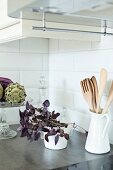 House plant next to white china jug of wooden spoons and bowl of artichokes on surface in corner