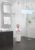 Toddler wearing dressing gown in front of glass screen of floor-level shower; heated towel rack in background