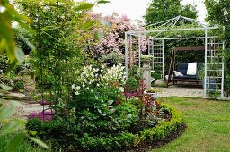 Round bed edged by low hedge in garden in front of garden swing in metal pergola