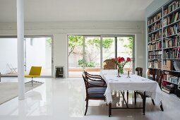 Table with white tablecloth and armchairs opposite bookcase in loft apartment: terrace doors with view into courtyard in background